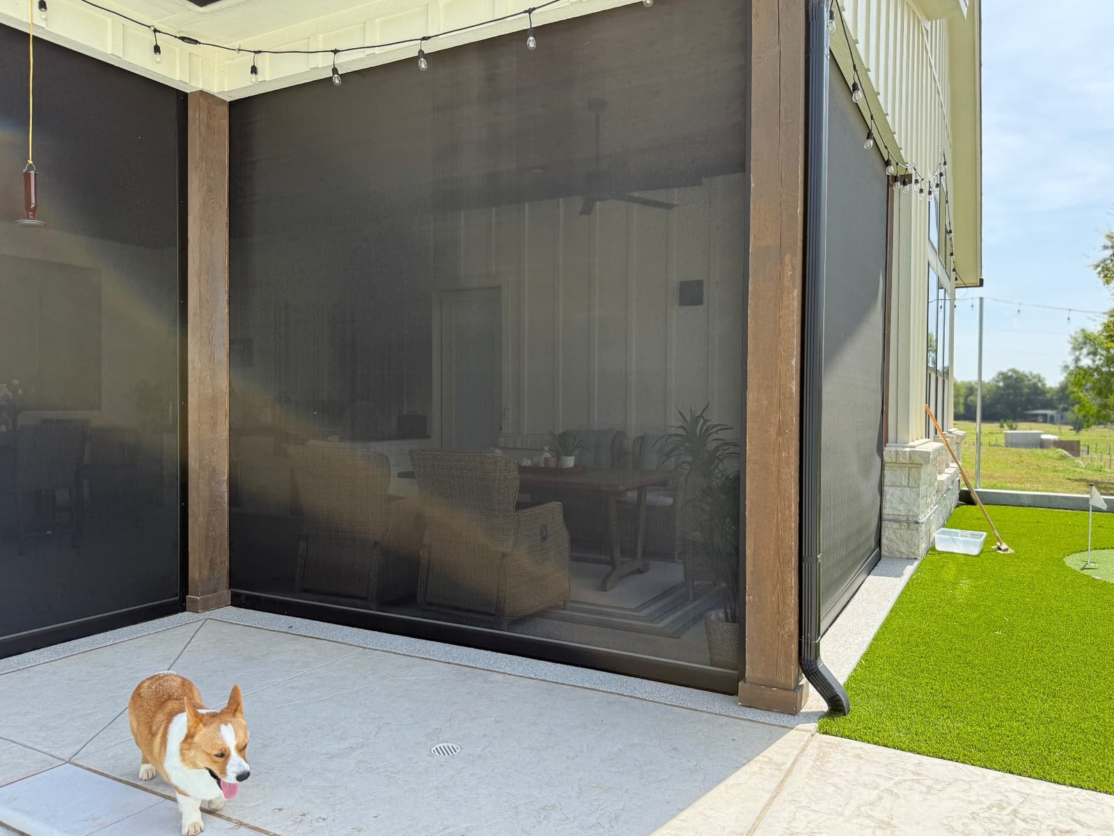 A covered patio with dark retractable screens, string lights overhead, a corgi on the stone floor in the foreground, and a putting green and open lawn beyond.