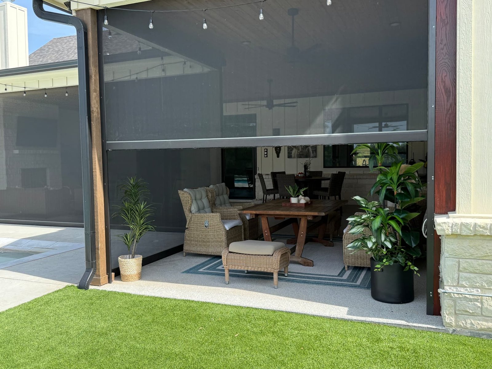 A covered patio lounge with retractable screens, woven seating, potted plants, and a view toward the pool and home.