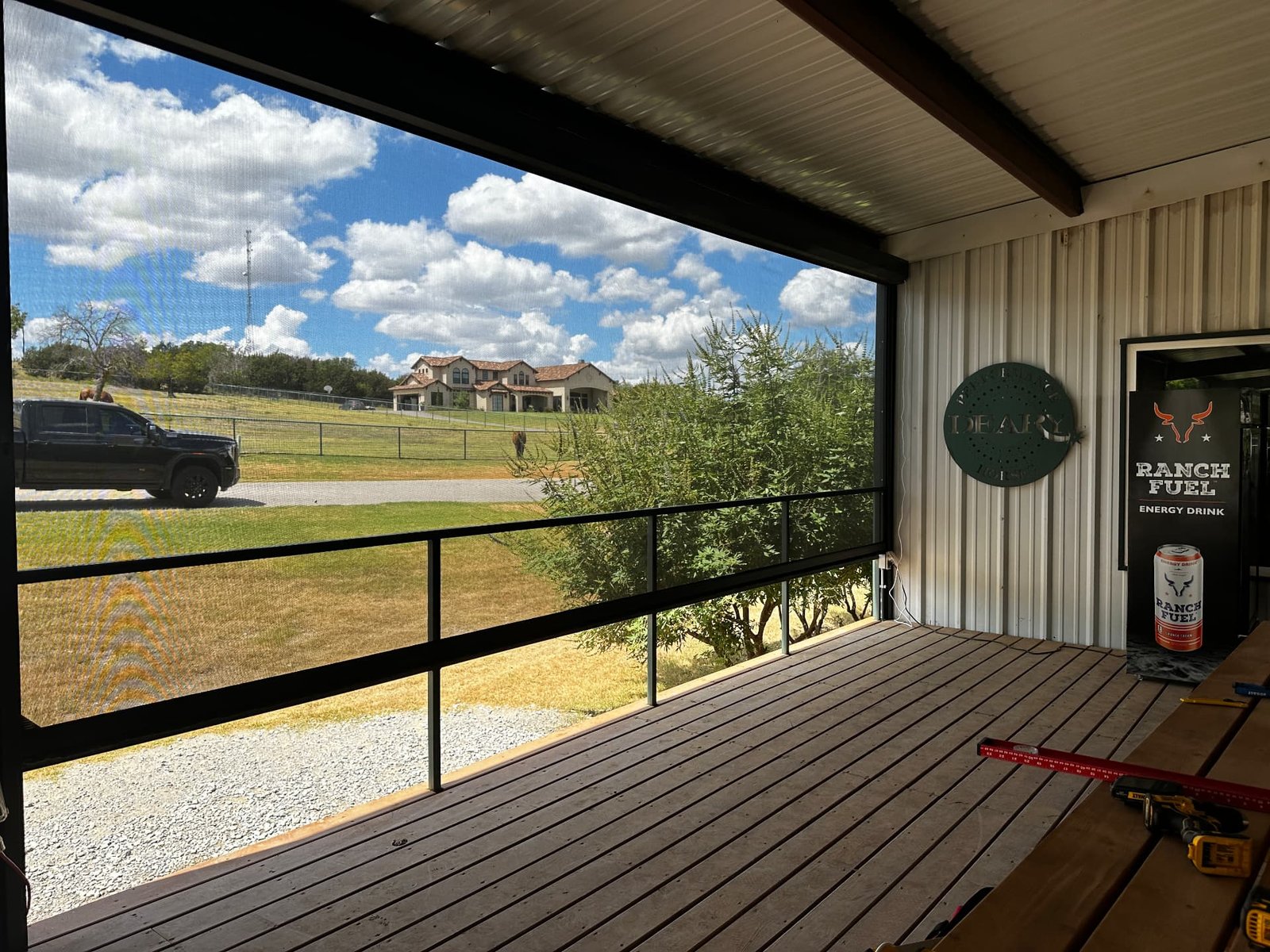 A covered ranch deck with railing overlooking pasture, truck, horse, and distant home under a wide Texas sky.