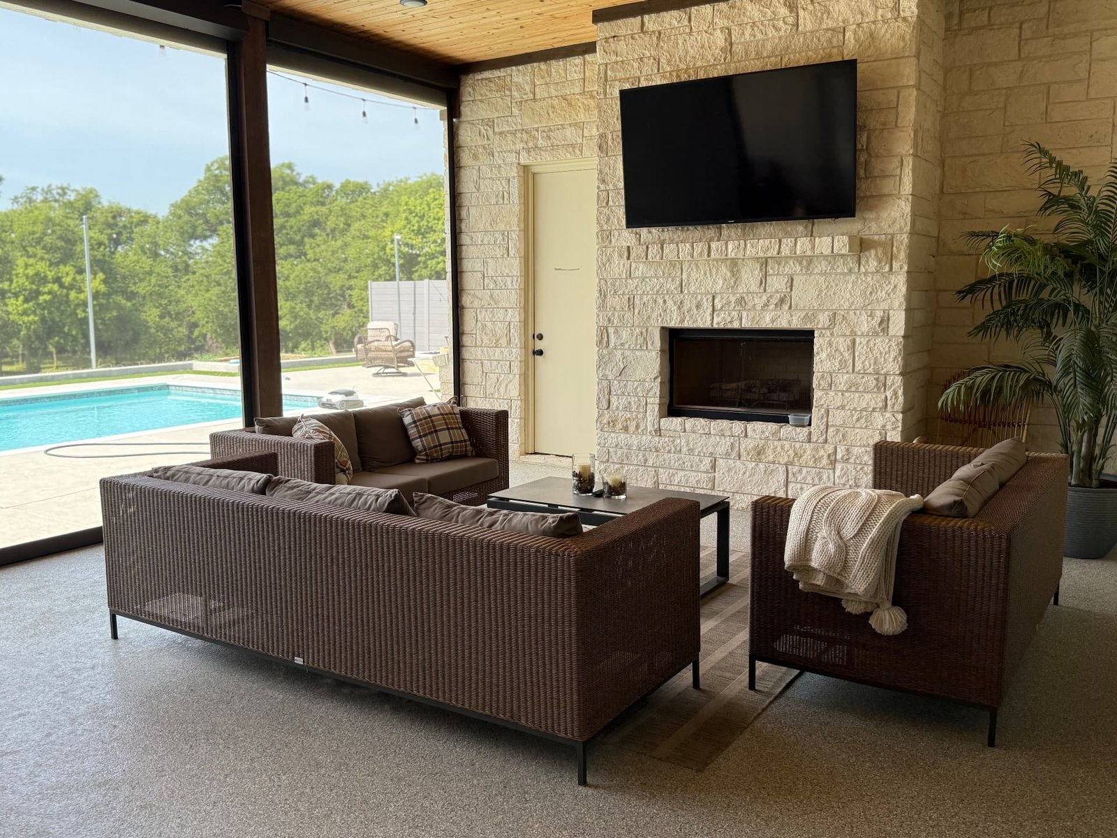 A covered patio lounge with woven seating, a stone fireplace, wall-mounted television, and retractable screens opening to the pool.