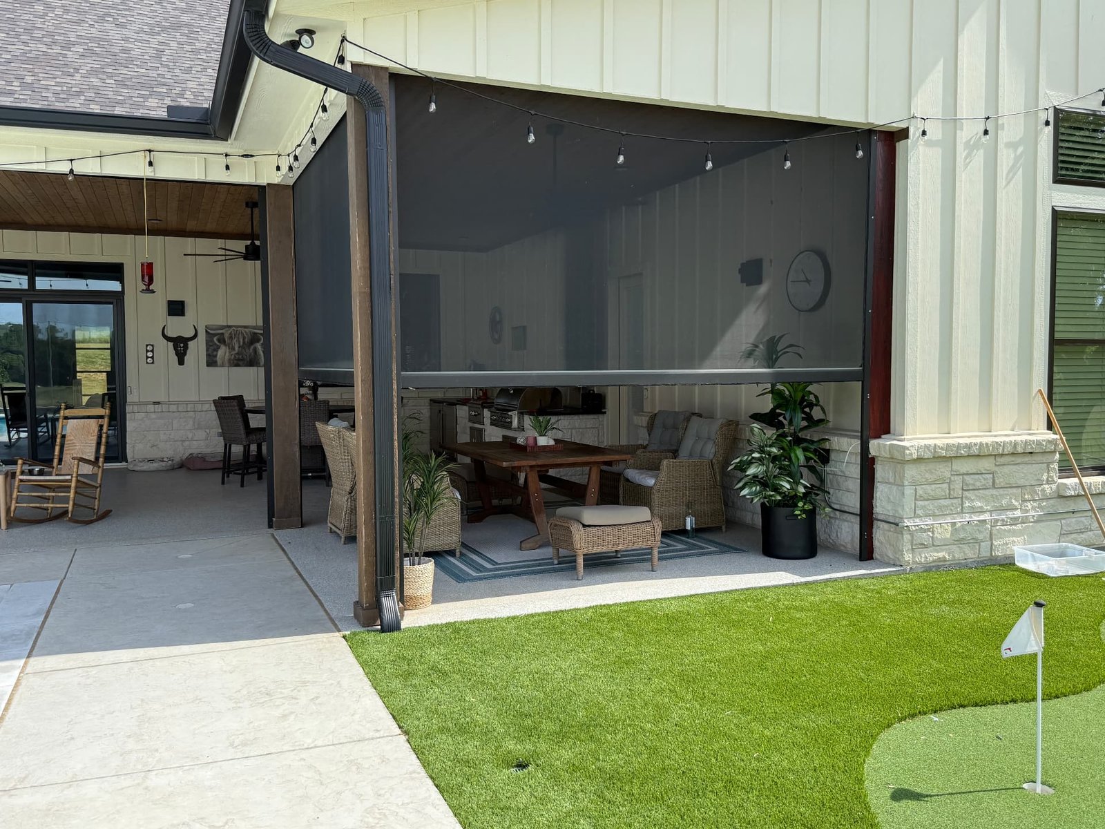 A covered patio dining and lounge area with dark retractable screens, string lights, woven seating, a wooden table, and a putting green and lawn in the foreground.