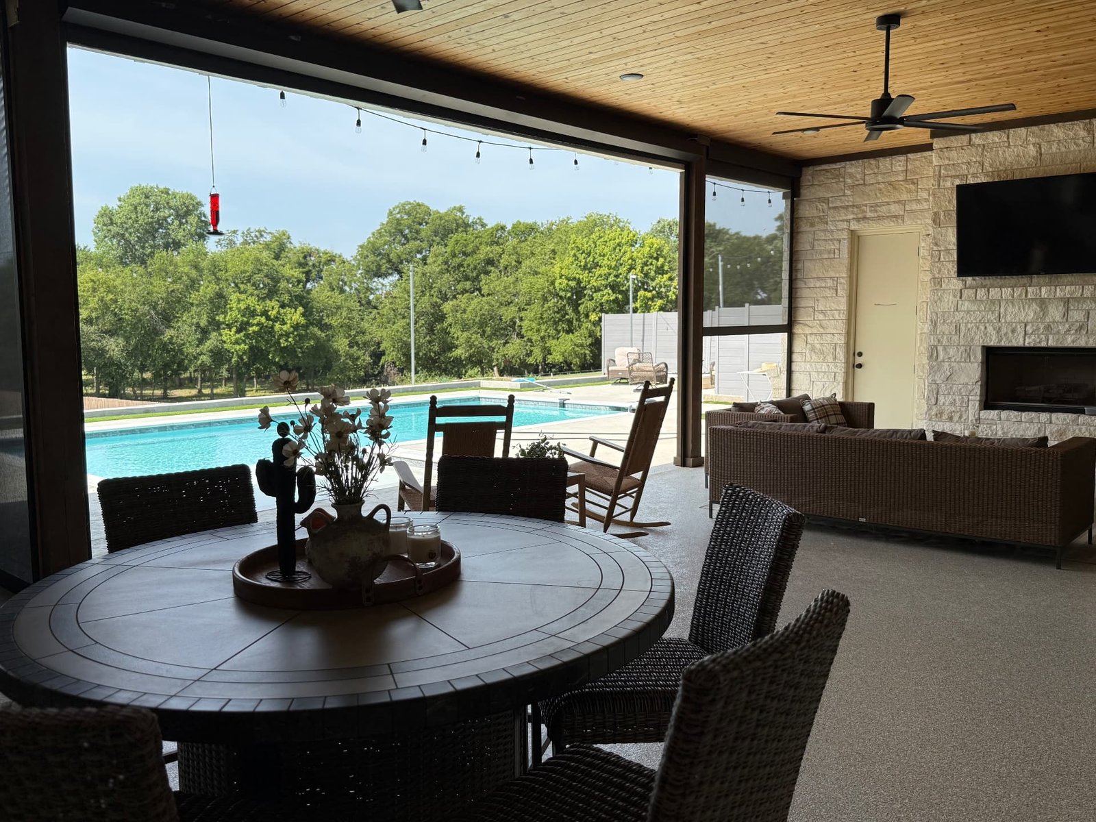 A covered patio dining and lounge space with a round table, woven seating, a stone fireplace, and retractable screens opening to the pool and landscape.